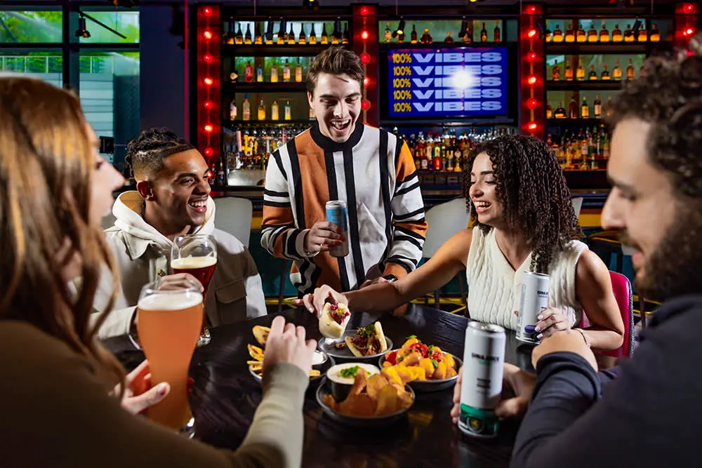 Three men and two women sharing and enjoying food and drinks at a table in Puttshack
