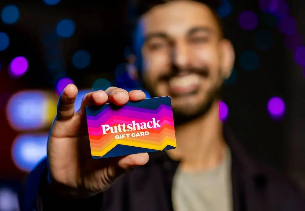 a young man smiling while holding up a puttshack gift card